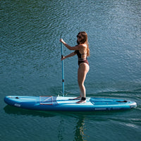 Femme debout vue profil sur SUP gonflable Selka 10' bleu-vert Aquadesign, lac vert en arrière-plan